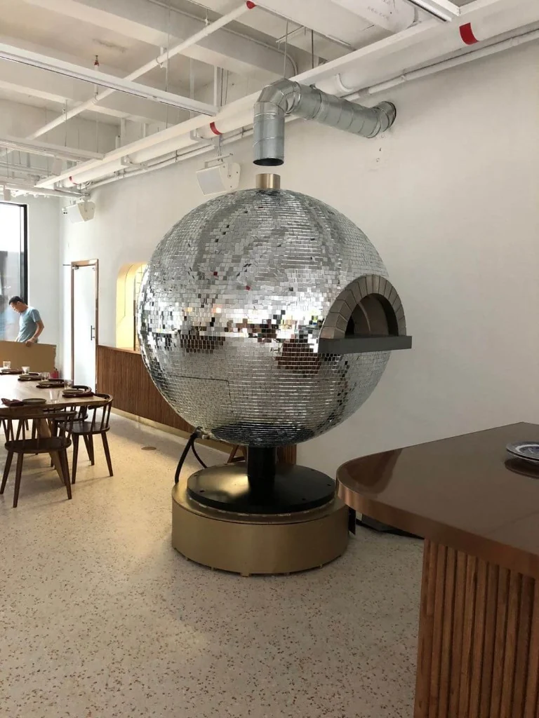 Custom mirror mosaic spherical pizza oven reflecting the dining room, with stone-trimmed arch and brass base in a bright restaurant interior