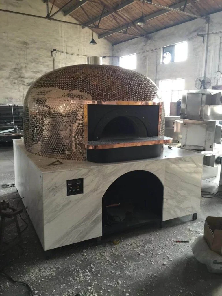 Large dome outdoor pizza oven with copper honeycomb mosaic tiles, white marble-look base, arched baking opening, and digital control panel in a factory workshop