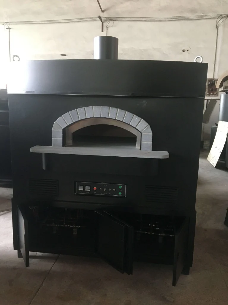 Large matte black dome outdoor pizza oven with stone-trimmed arch, stone landing shelf, dual digital control panel, front vents, and open lower cabinet showing gas plumbing in a factory setting