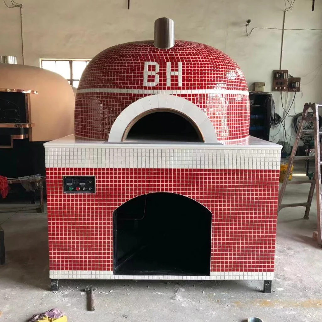 Custom red and white mosaic dome outdoor pizza oven with BH logo tile, stainless chimney, digital control panel on the base, and workshop background