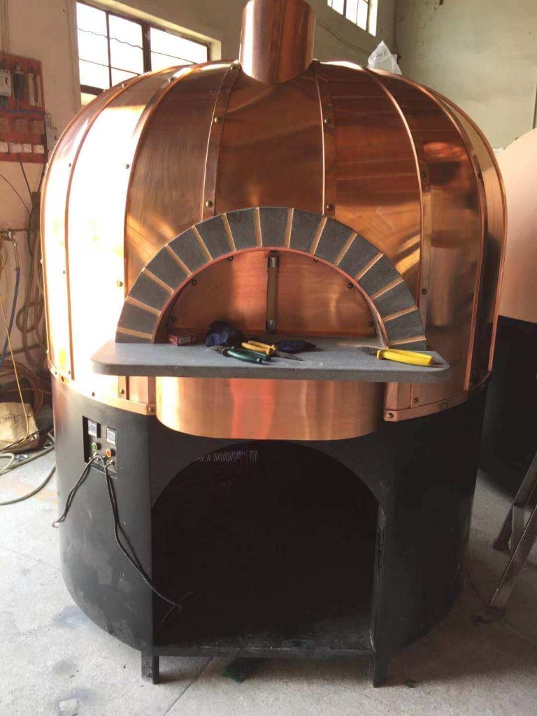 Custom copper dome pizza oven with riveted panels, stone landing, and digital controls on a black base in a factory workshop