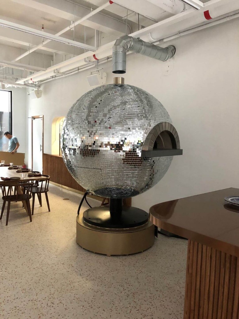 Custom mirror mosaic spherical pizza oven reflecting the dining room, with stone-trimmed arch and brass base in a bright restaurant interior