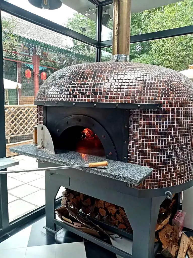Commercial wood-fired pizza oven with bronze mosaic dome, stone shelf, and firewood storage on a metal stand