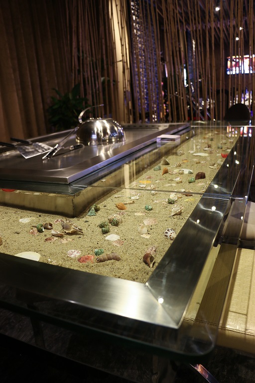 Close-up of recessed glass-topped sand and shell inlay in a brushed stainless frame beside a teppanyaki cooktop