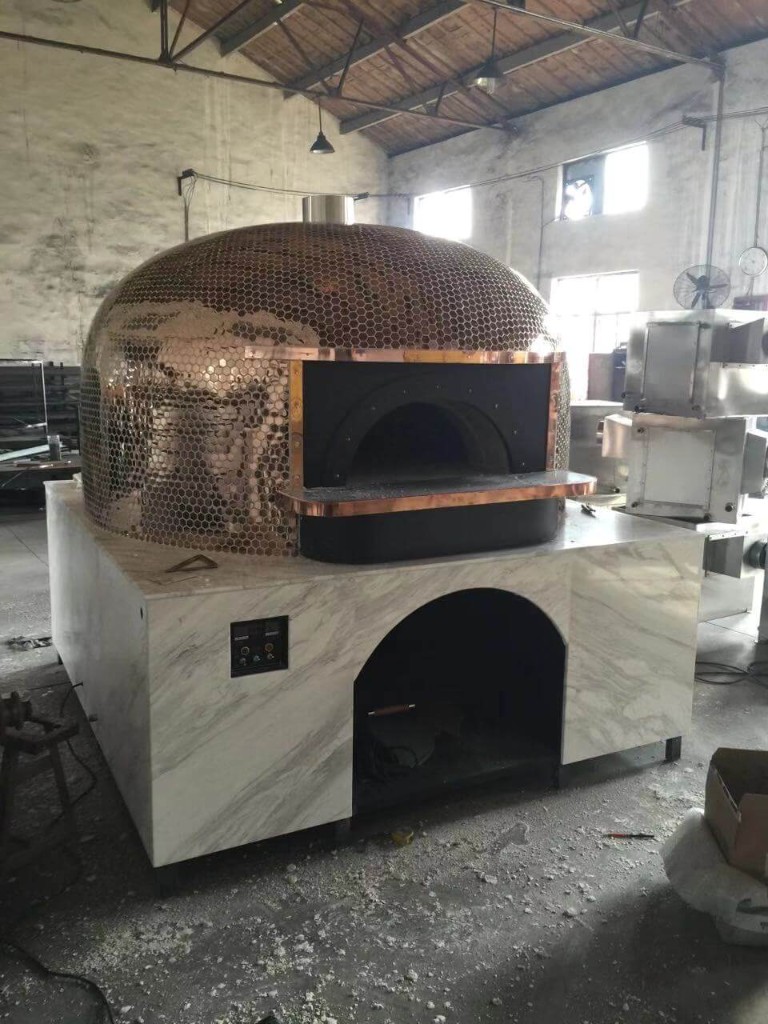 Large dome outdoor pizza oven with copper honeycomb mosaic tiles, white marble-look base, arched baking opening, and digital control panel in a factory workshop