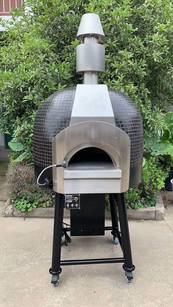 Portable dark mosaic dome outdoor pizza oven on a black wheeled stand with stainless arch, chimney stack, digital controller on the leg, and garden greenery behind