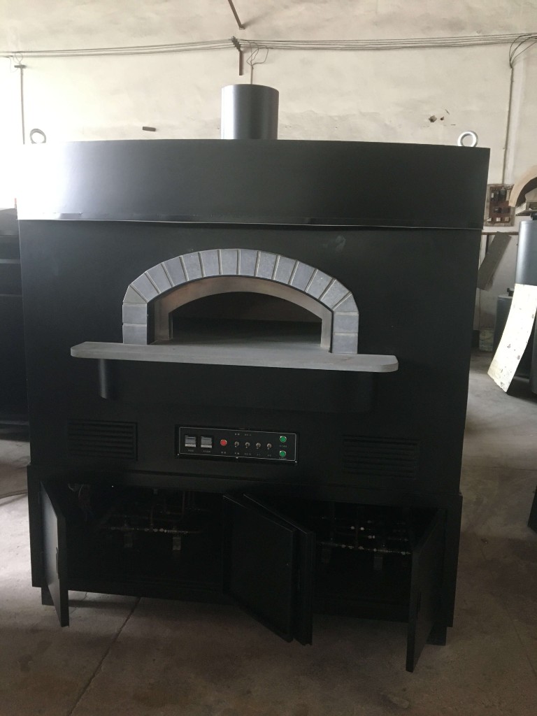 Large matte black dome outdoor pizza oven with stone-trimmed arch, stone landing shelf, dual digital control panel, front vents, and open lower cabinet showing gas plumbing in a factory setting
