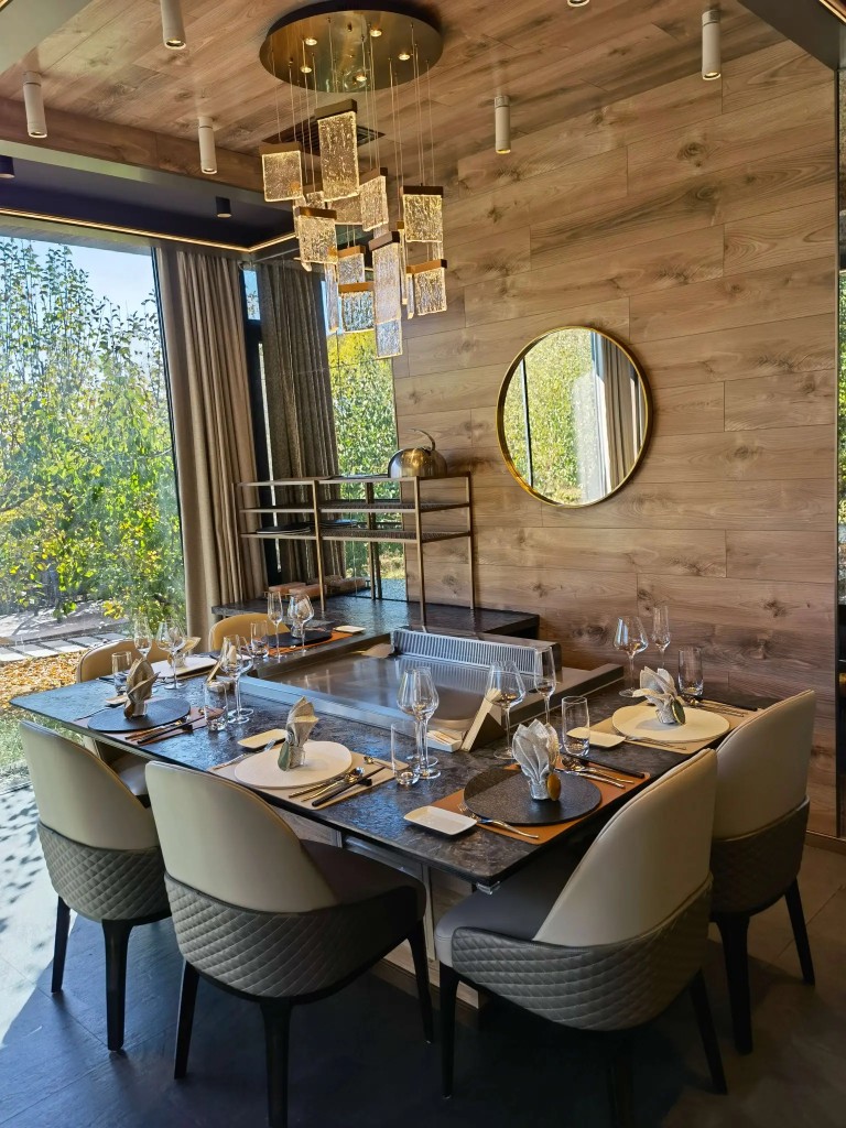 High-end residential dining room with built-in teppanyaki grill, chandelier, wood walls, and garden view