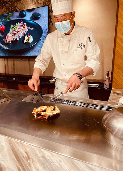 Male teppanyaki chef in white uniform cooking on a stainless steel grill with screen behind